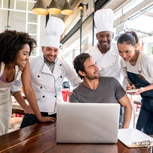 man sitting at a table with a laptop and four restaurant workers standing around him