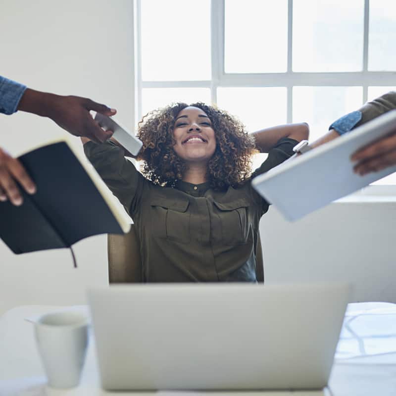 woman leaning back at her desk with arms back, eyes closed, smiling while coworkers are handing things to her