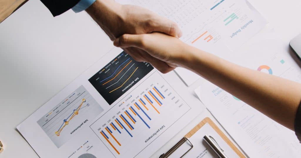 An overhead shot of a desk covered with charts, graphs, and financial reports. Two people shake hands over the desk.