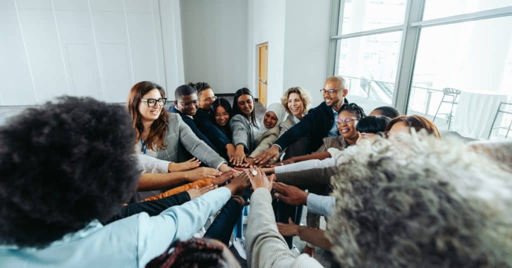 A large, diverse team of professionals sits in a circle. They all smile and put one hand into the middle of the room.