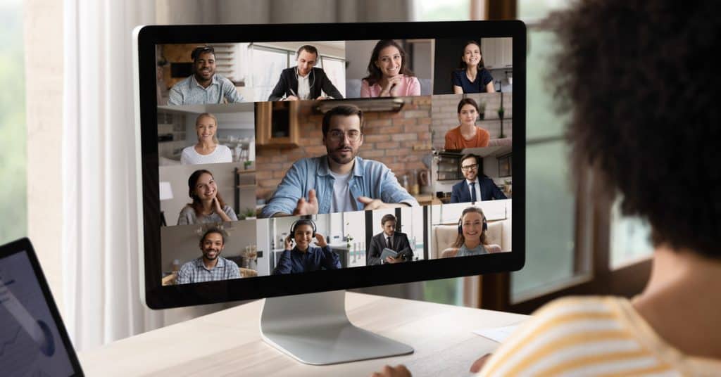 A young woman sits at a desk in her home office. She faces her computer screen and joins a multi-person video call.